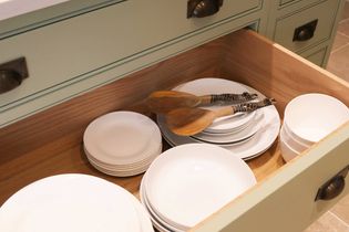 Open kitchen drawer with neatly stacked white plates, bowls, and wooden spoons. The drawer has a pale green exterior, creating an organized, calm vibe.