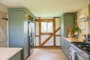 A rustic kitchen with green cabinetry, exposed wooden beams, a stainless steel refrigerator, and a window letting in natural light. Cozy and inviting atmosphere.