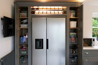 A sleek, shaker kitchen with a large silver fridge centered between open pantry cabinets. The top features a wine rack, and a microwave is on the right.
