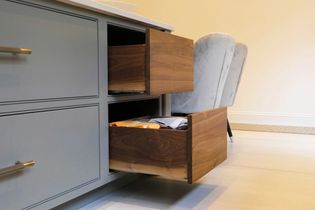 Open wooden kitchen drawers in a modern setting, showing neatly arranged snacks. Softly lit environment with light grey cabinetry and plush chairs.