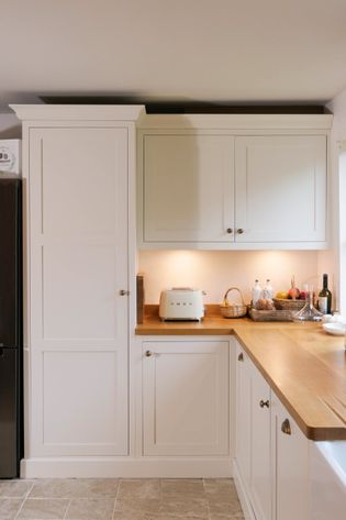 Shaker kitchen corner with white cabinets and wood countertops. A toaster, basket, and bottles sit under warm under-cabinet lighting.