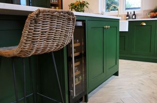 Cozy kitchen with green cabinets and brass knobs. Features a wicker stool, small wine cooler, white countertops, and fresh flowers on the counter.
