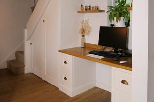 Shaker style home office under stairs with wood desk, white drawers, computer, keyboard, mouse, shelf with plants, and soft lighting for a warm feel.