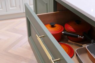 Open kitchen drawer with brass handle reveals orange and red pots with black knobs. The drawer is beneath a white countertop, on a light wood floor.