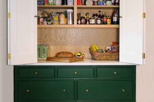 Open kitchen pantry with white doors, showcasing organized shelves of food items, a loaf of bread on a cutting board, and a basket with bananas on a green dresser.