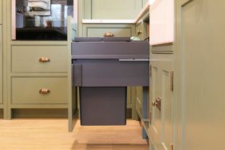 A sleek, shaker kitchen with green cabinets and brass handles. A pull-out drawer reveals a dark grey, recycling bin system.
