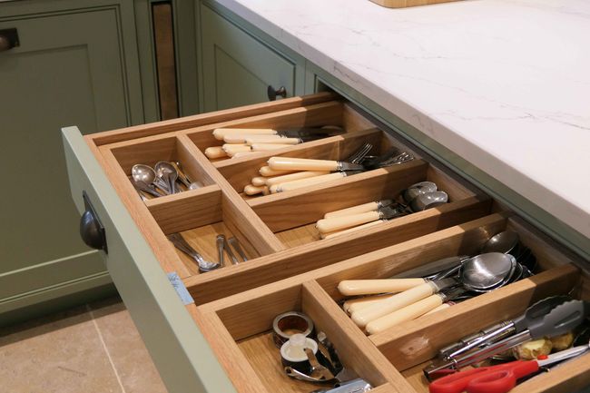 Open kitchen drawer with organized cutlery in wooden compartments. Cream-handled knives, forks, spoons, and utensils against a marble countertop.