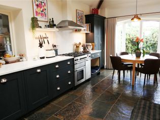 Spacious kitchen with dark cabinetry, a modern range, and a sleek hood. A round table with chairs overlooks a garden through large windows. Earthy tones create a cozy ambiance.