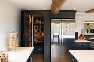 Shaker kitchen with sleek stainless steel fridge, open pantry filled with food items, dark cabinets, exposed wooden beam, and a bright, clean ambiance.