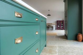 A shaker kitchen features green cabinets with brass handles, stone tile floor, and a purple cupboard in the background, creating a elegant atmosphere.