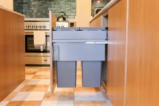 Two grey pull-out bins beneath a kitchen counter, with an oven, kettle, and tiled backsplash in the background.