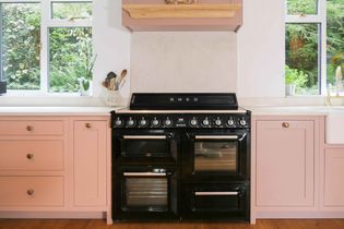 A sleek black Smeg oven is set between pastel pink cabinets in a bright kitchen. Two windows with lush green views add a fresh, serene feel.