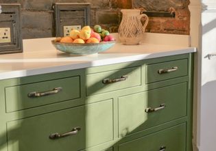 Freestanding green Shaker dresser within an open fireplace, with multiple drawers, a fruit bowl and ceramic jug is on top of the white quartz worktop.