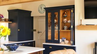 A cozy kitchen with navy cabinets and wooden shelves showcasing colorful ceramics. A sunflower bouquet adds cheer, while a wall clock shows the time.