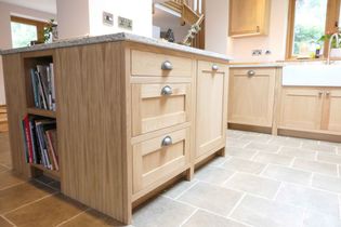 Shaker kitchen with oak cabinetry, silver handles, and a granite countertop. Island is the focus of this image with open book shelving and an open front sink is behind.