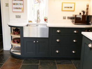 Shaker kitchen with slate floor, dark cabinetry, and stainless steel sink. Open shelves display colorful tins. A microwave sits on the counter nearby.