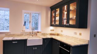Spacious shaker kitchen with navy blue cabinets and granite worktop, featuring a farmhouse sink, glass-door cupboards, and soft, warm lighting.