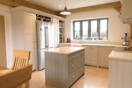 Bright country style Shaker kitchen with a small central island containing three drawers, exposed wooden beams and a large window above the sink.