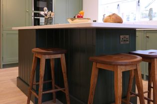 Shaker kitchen with a dark green island, white worktop, and wooden stools. Light wood floor, green cabinetry, bread, and utensils provide a cosy, rustic feel.