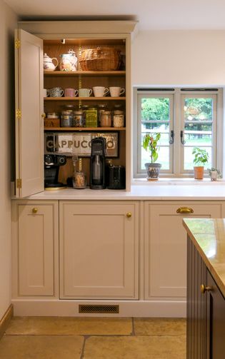 Open Shaker kitchen cupboard stocked with teapots, cups, jars, and coffee appliances beside a window with potted plants.