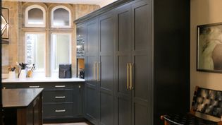 Shaker kitchen with dark cabinets and brass handles, white countertop with utensils, two arched windows, and a painting on the wall. Cozy ambiance.