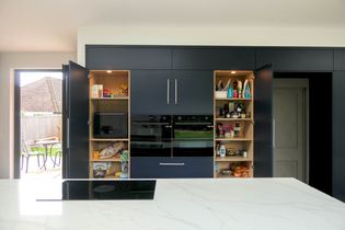 Modern kitchen with open dark cabinets revealing shelves of groceries and appliances. A large island with a stovetop stands in the foreground, adjacent to a sunlit patio.