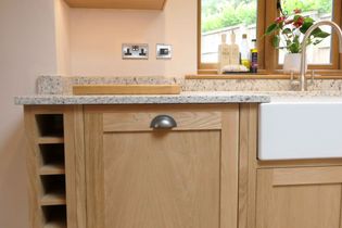 Wooden kitchen cabinetry with a granite countertop, featuring a metal handle and integrated wine rack. A farmhouse sink with a tap is nearby, with bottles and a vase on the windowsill, creating a cozy atmosphere.