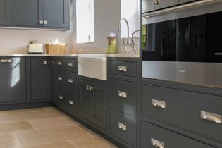 Shaker kitchen with dark grey cabinets, silver handles, and a sleek built-in oven. A white farmhouse sink sits under a bright window.