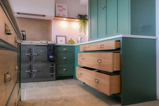 Shaker kitchen with green cabinets, open wood drawers, and a grey stove. Warm lighting highlights a framed picture and plant on the counter.