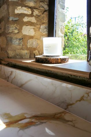 Rustic stone wall and window with a lit candle on a wooden coaster, natural sunlight comes in. Marble look countertop in the foreground.