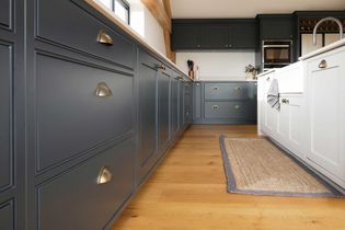Shaker kitchen interior with dark grey cabinets and brass handles. Light wood flooring and a woven rug add warmth. White walls and ample lighting create a clean, cozy feel.