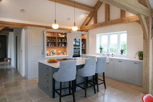 Spacious shaker kitchen with light grey cabinets, central dark grey island with three barstools, pendant lights, wooden beams, and a large window with plants.