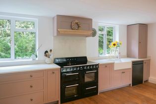 A cozy kitchen with pink cabinets, black oven, and farmhouse sink. Sunflowers in a vase sit by the window. Wood floor adds warmth. Serene ambiance.