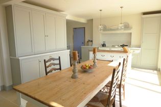 Bright kitchen with a wooden table, four chairs, and a bowl of fruit. Cream-coloured cabinets, pendant lights, and large windows enhancing natural light.
