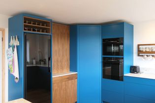 A modern kitchen with blue and wood cabinets, a sleek black fridge, two built-in ovens, and a countertop with appliances. A wine rack and a hanging towel add detail.