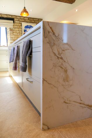 Kitchen waterfall island, grey drawers, silver handles, and kitchen towels hanging on handles; stone wall and window in the background.