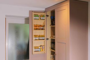 Tall shaker kitchen cabinet with a door open, revealing spice rack filled with jars on the inside. Adjacent shelves hold various pantry items. Bright lighting.