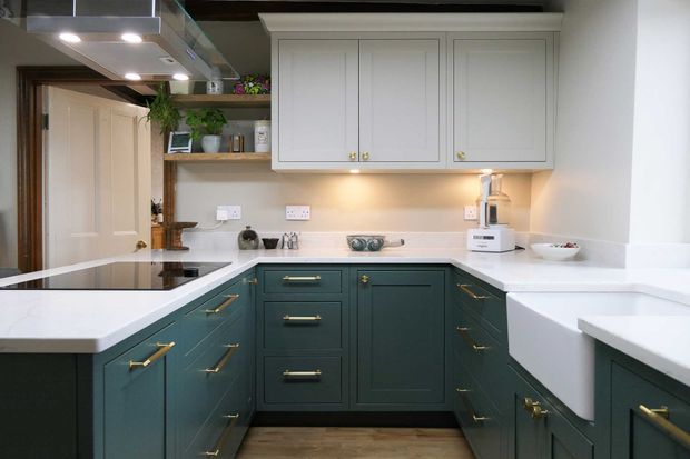 U-shaper Shaker kitchen with dark teal cabinets around the perimeter and white cabinets above. Brass hardware is used throughout, with a farmhouse sink and open wooden shelving to the left of the white cabinets.