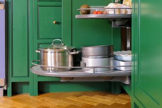 Green kitchen cabinet with a pull out corner shelf holds pots, pans, and dishes. Items include a stainless steel pot and baking tins, on a wooden floor.