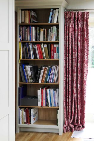 A bookshelf of 6 shelves filled with various books, next to a window adorned with red patterned curtains. 