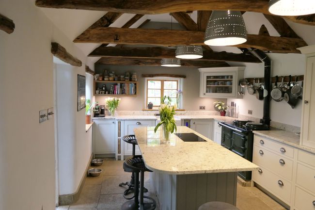 Rustic shaker kitchen with exposed wooden beams and stone floor. A central island with stools, hanging pans, and fresh flowers create a warm, inviting atmosphere.