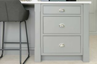 A shaker kitchen features a grey cabinet with three drawers and shiny silver handles. Beside it, a sleek, cushioned black bar stool is partially visible.