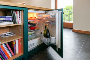Open under-counter fridge in the kitchen island, stocked with vegetables, bottles, and containers. Nearby, a bookshelf holds colorful cookbooks. Bright, inviting scene.