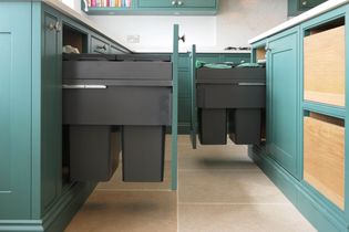 Pull-out kitchen cabinets with green and wood panels reveal black recycling bins on a tiled floor. Bright, organized, and modern kitchen space.