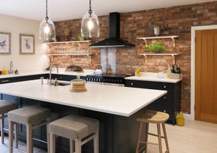 Shaker kitchen with brick wall, large white island, and grey stools. Pendant lights hang above, and shelves display plants and glassware, creating a cozy vibe.
