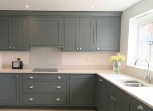 A shaker kitchen with grey cabinets and silver handles, featuring a quartz worktop with a toaster and a vase of yellow flowers by a large window. Bright and tidy atmosphere.