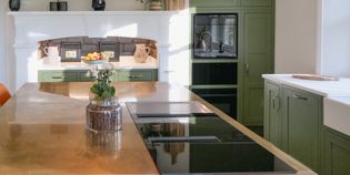 A close up of an induction hob on the brass island with a green plant next to it, ovens and dresser within an open fireplace in the background.