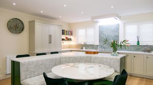 A Shaker kitchen featuring a circular dining table and a built-in seating area, with light-coloured cabinets and large windows for natural light