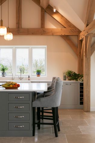 Spacious kitchen with exposed wooden beams, large windows, and beige walls. Features a dark grey island with grey chairs, pendant lights, and plants by the sink.