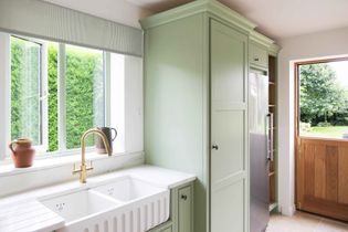 Bright shaker kitchen with pastel green cabinets, a white double sink, and brass tap. Light streams through a large window, revealing a lush garden outside.